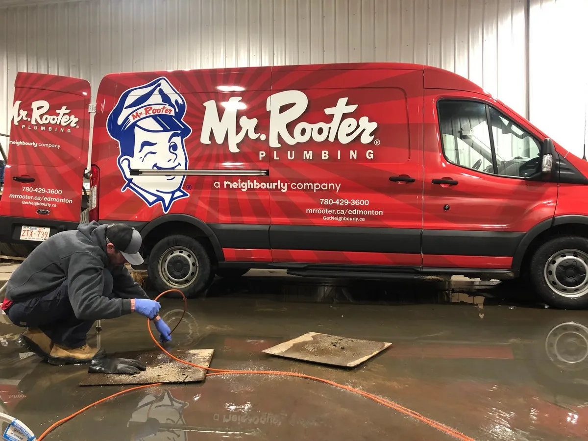 A plumber in Sherwood Park lowering a sewer scope into a floor drain with a large, branded Mr. Rooter Plumbing van visible in the background.