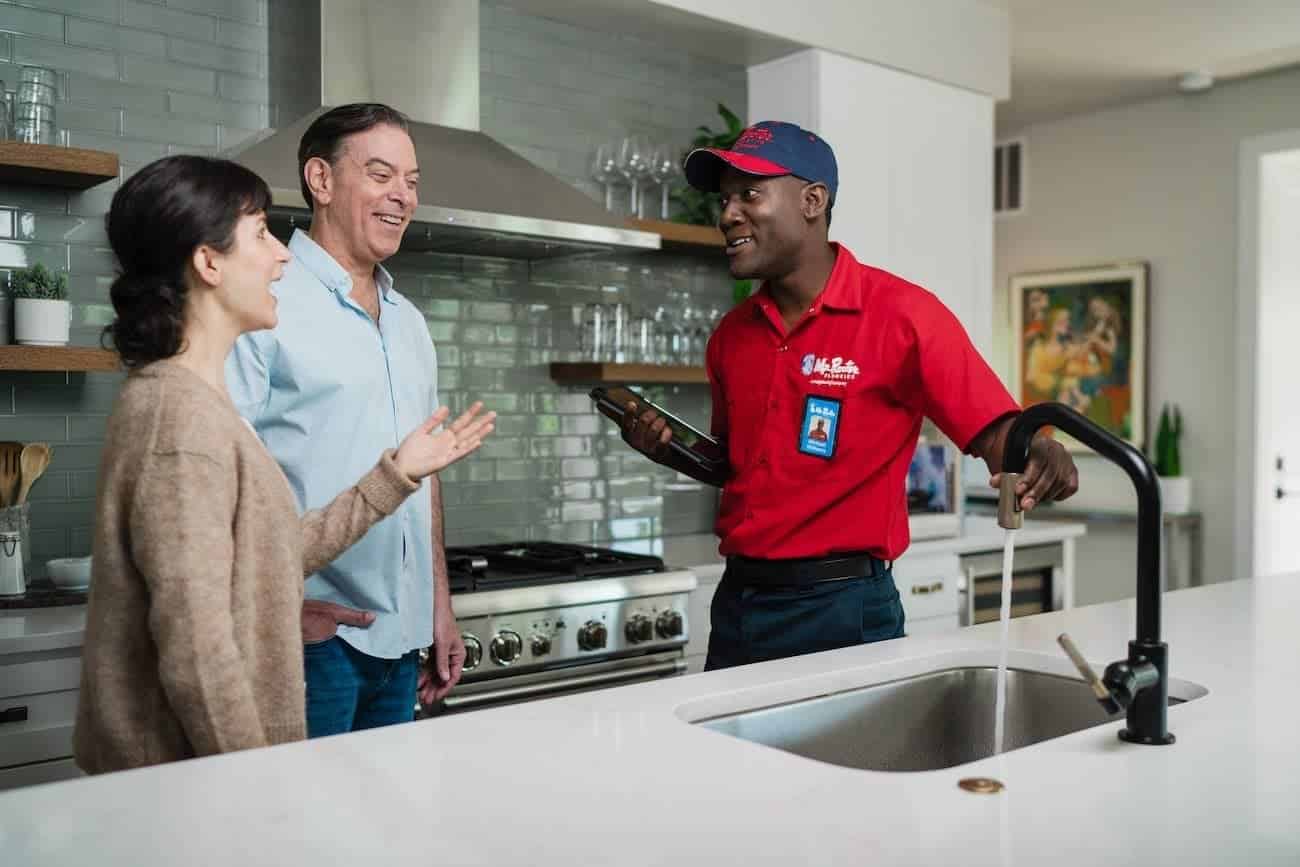 A uniformed Mr. Rooter professional discussing services with customers in their kitchen